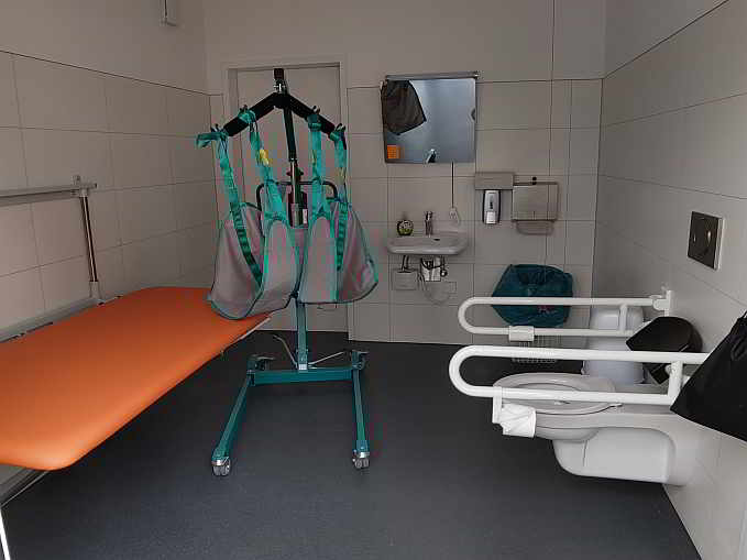 Die 8,8 qm große Rollstuhl-Toilette ist zusätzlich ausgestattet mit einer höhenverstellbaren Wandklappliege für Erwachsene, einem mobilen Patientenlifter (Hebetuch vorhanden) sowie einem luftdichten Windeleimer.